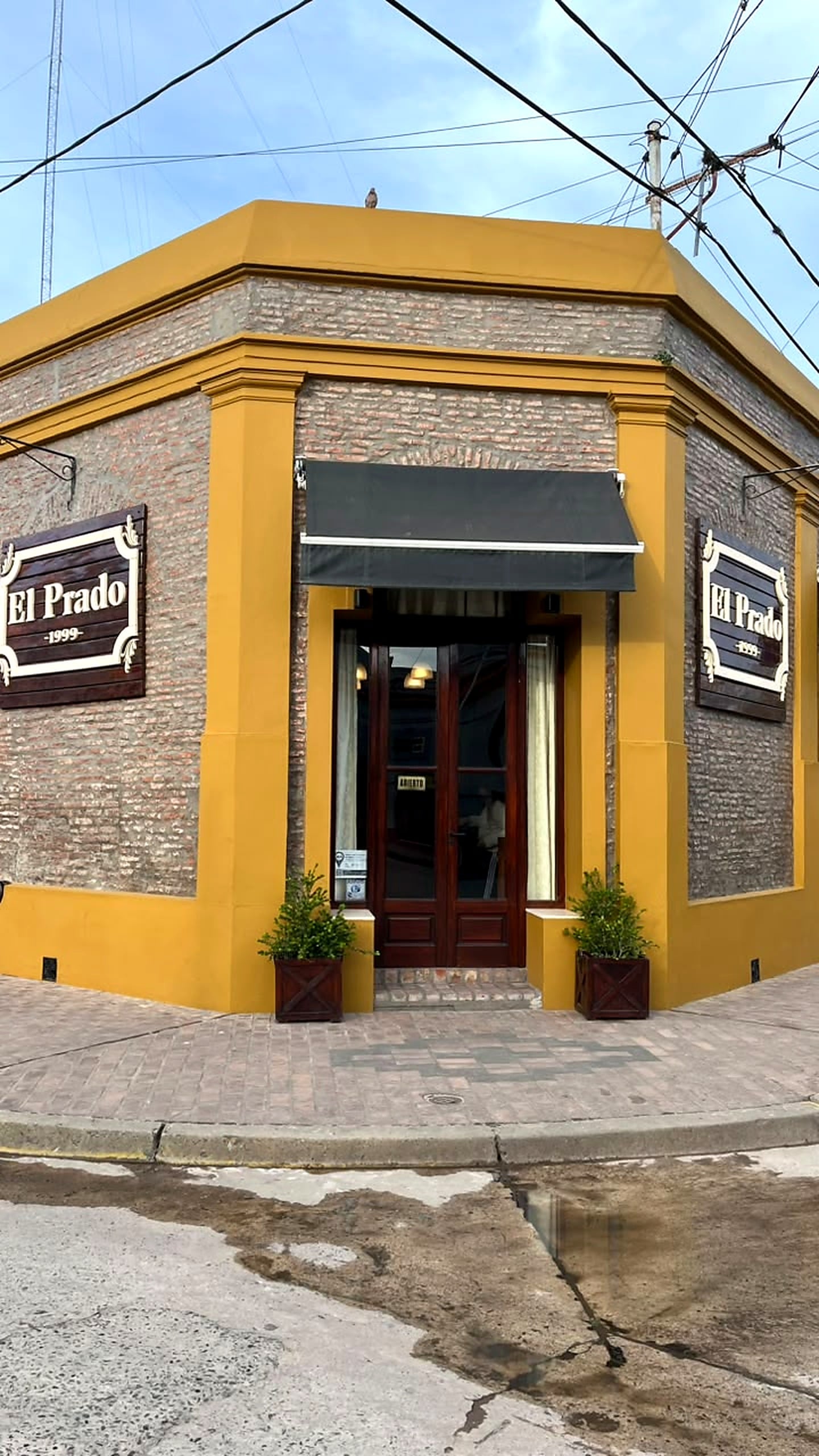 Fachada de El Prado: edificio de esquina color mostaza con ladrillo a la vista, carteles de madera con el nombre y la puerta de madera en la ochava.