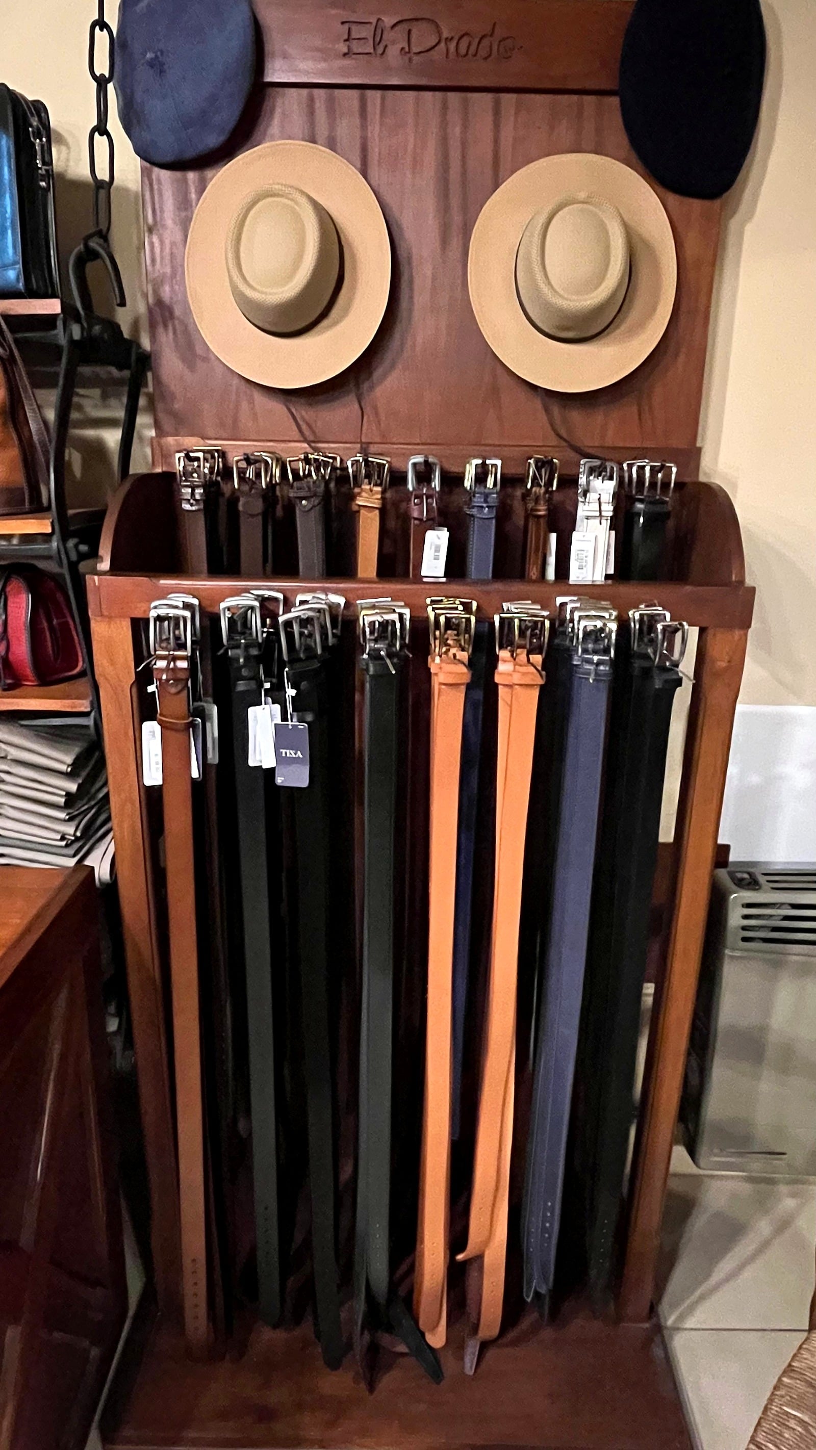 Exhibidor de madera con el nombre El Prado tallado, dos sombreros de ala, boinas y cinturones colgados.